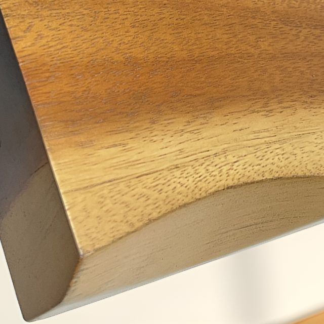 Close-up of the sculpted underside of a suar shelf in sunlight, showing the smooth clear-coated grain